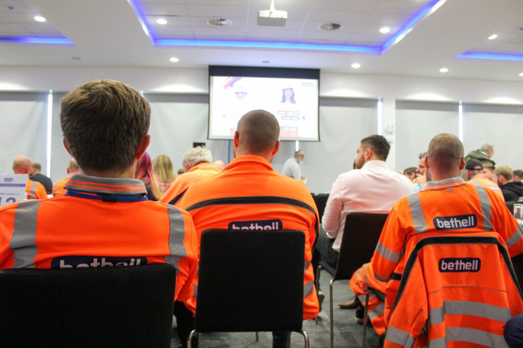 A group of operatives in orange PPE sat in chairs at a large conference